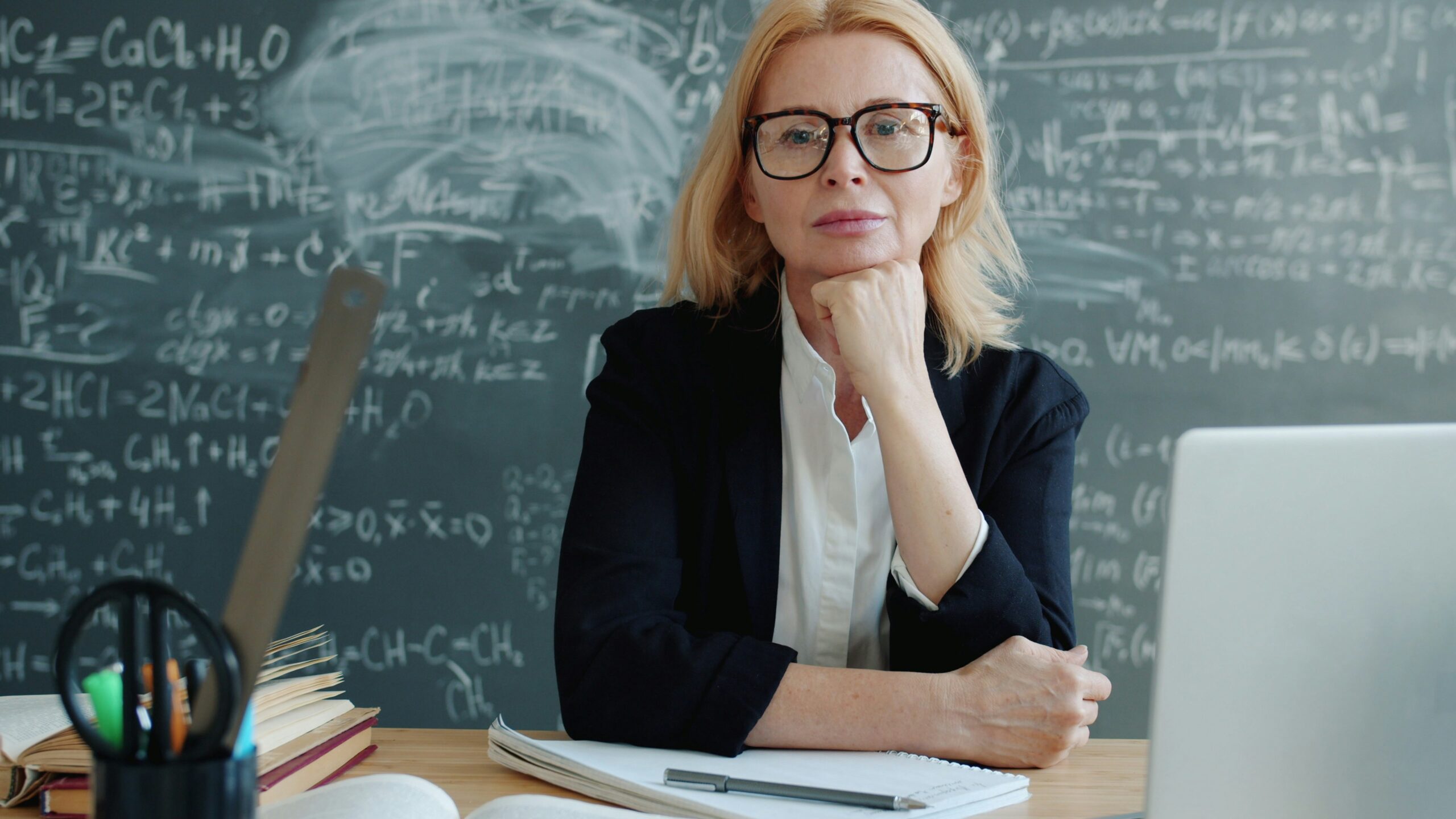 Serious adult woman is sitting at desk in university classroom looking at camera wearing suit and glasses. Profession, lifestyle and education concept.