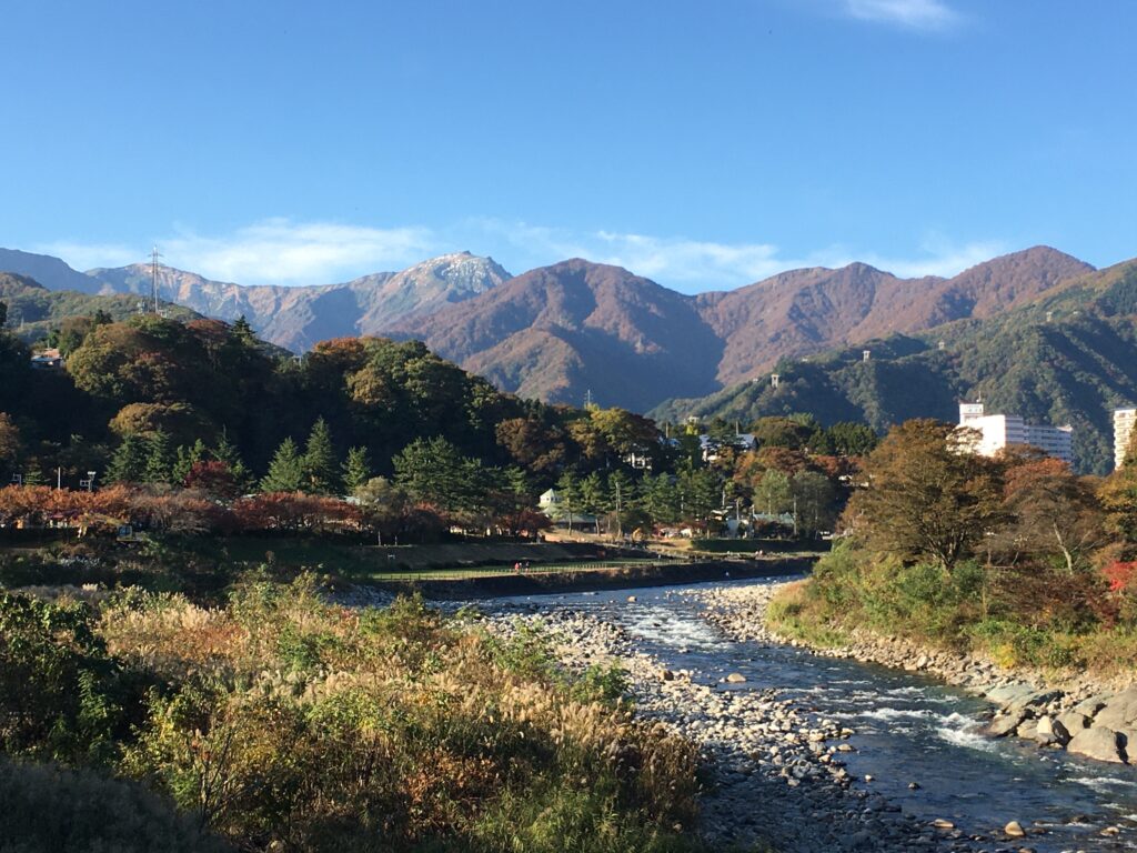 A shallow, rocky river flows toward the viewer between grassy banks, with a small town nestled among autumn-coloured trees and forested mountains rising behind under a blue sky. The snow-traced peak of Tanigawadake is visible in the distance.